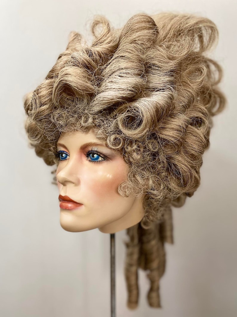 K&ouml;nnte beinhalten: Eine blonde Per&uuml;cke, die zu einer gro&szlig;en, aufwendigen Hochsteckfrisur mit Locken und Ringellocken gestylt ist. Die Per&uuml;cke wird auf einem Mannequin-Kopf pr&auml;sentiert.