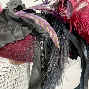 May include: A burgundy felt hat with black feathers, black ribbon, and black netting. The hat is decorated with sequins and has a purple feather with gold accents.