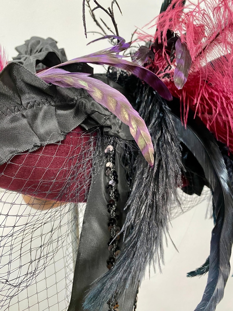May include: A burgundy felt hat with black feathers, black ribbon, and black netting. The hat is decorated with sequins and has a purple feather with gold accents.