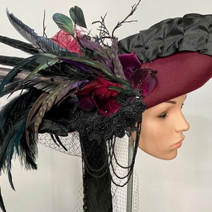 May include: A burgundy felt hat with a black satin ruffle and a large black feather plume. The hat is adorned with black velvet flowers, black beads, and black lace.