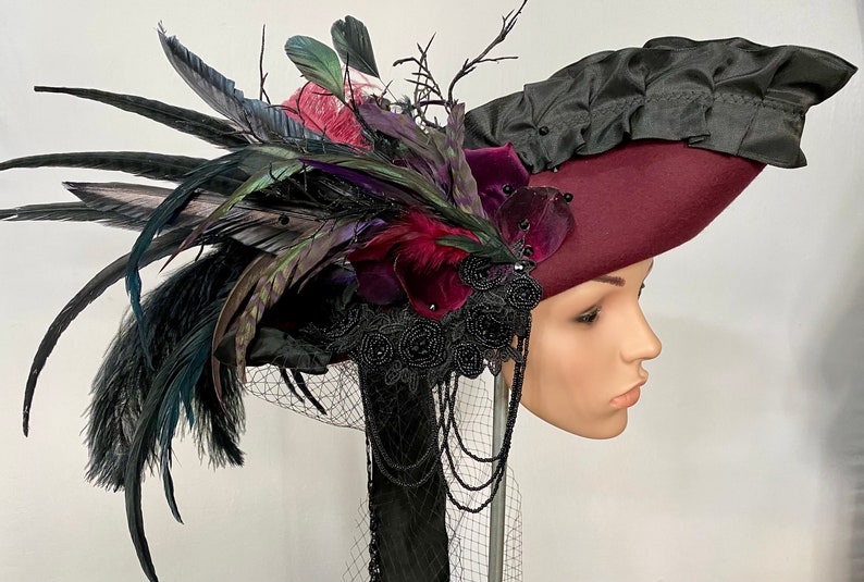 May include: A burgundy felt hat with a black satin ruffle and a large black feather plume. The hat is adorned with black velvet flowers, black beads, and black lace.
