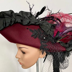 May include: A burgundy felt pirate hat with a black ruffle trim, black lace embellishments, and purple and burgundy feathers.
