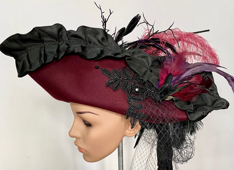 May include: A burgundy felt pirate hat with a black ruffle trim, black lace embellishments, and purple and burgundy feathers.