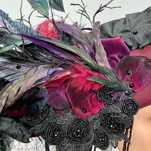 May include: A burgundy velvet hat with black feathers, black lace, and black beads. The hat is decorated with a variety of feathers, including black, purple, and green feathers.