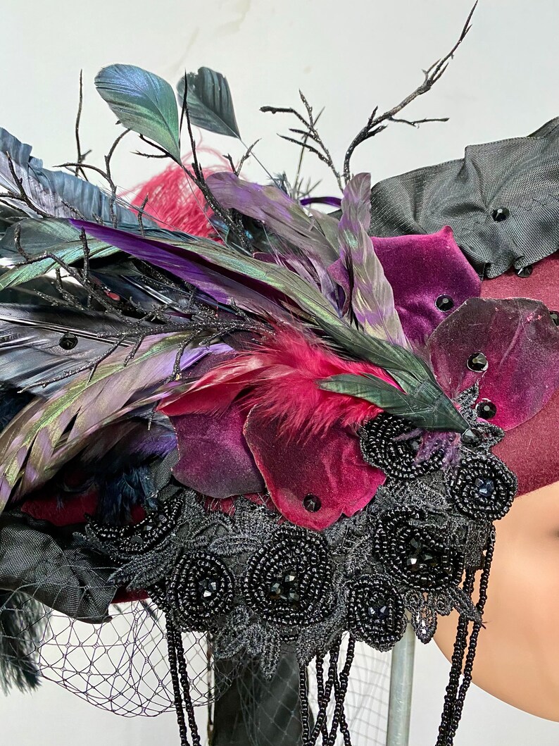 May include: A burgundy velvet hat with black feathers, black lace, and black beads. The hat is decorated with a variety of feathers, including black, purple, and green feathers.