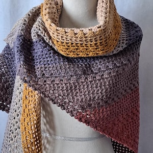 May include: A crocheted shawl with a gradient of colors, including brown, purple, orange, and red. The shawl is draped over a white mannequin.