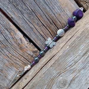 May include: A beaded bookmark with a silver clip. The bookmark features purple and black beads, a silver butterfly charm, and a decorative silver disc. The clip is attached to a strand of beads and purple stones.