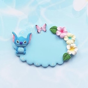 May include: A light blue ceramic name tag with a scalloped edge, featuring a blue Stitch character and a pink butterfly. The tag is decorated with a floral garland of pink, white and yellow flowers.