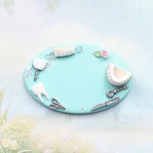 Può includere: Un elemento decorativo ovale azzurro con strumenti odontoiatrici in miniatura e un dente. Il pezzo include un calco dentale, un dente, forbici, un bisturi e una piccola rosa rosa. Lo sfondo è una scena ad acquerello con uno stagno e fiori.