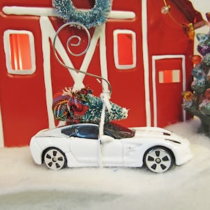 May include: A white toy car with a small Christmas tree tied to the roof. The car is in front of a red barn with white trim and a Christmas tree with lights.