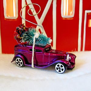 May include: A purple vintage car with a small Christmas tree tied to the roof. The car is a Christmas ornament.