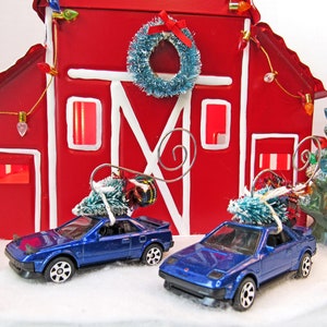 May include: Two blue toy cars with Christmas trees on top, parked in front of a red barn with white trim and a blue wreath on the door. The barn has a string of lights and a small Christmas tree next to it.