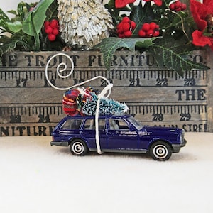 May include: A blue toy car with a small Christmas tree and a plaid blanket tied to the roof. The car is sitting on a white surface in front of a wooden box with a ruler and the text "The Trout Furniture Co. The Street" printed on it.