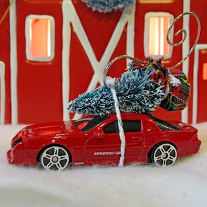 May include: A red toy car with white wheels and the text "IROC-Z" is decorated with a small blue Christmas tree and a plaid gift. The car is set against a red barn backdrop, creating a festive holiday scene.