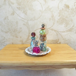 May include: A white tray with a ruffled edge holds three decorative bottles. The bottles have colorful glass stoppers and are filled with glitter and beads. The tray is on a light wood table.