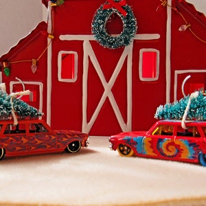 May include: Two red toy station wagons with Christmas trees on top, parked in front of a red barn with a white X on the door. The barn has a wreath on the top and lights hanging from the eaves.
