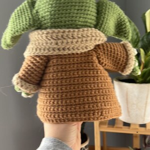 May include: A crocheted plush toy of a green character with brown clothing. The toy is being held by a hand.