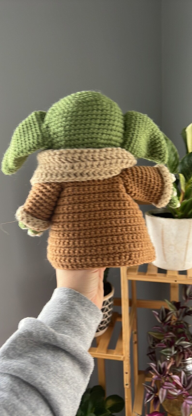 May include: A crocheted plush toy of a green character with brown clothing. The toy is being held by a hand.