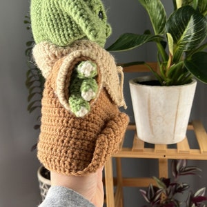 May include: A crocheted green and brown plush toy resembling a character from the Star Wars franchise. The toy is being held by a hand in a grey sweatshirt.