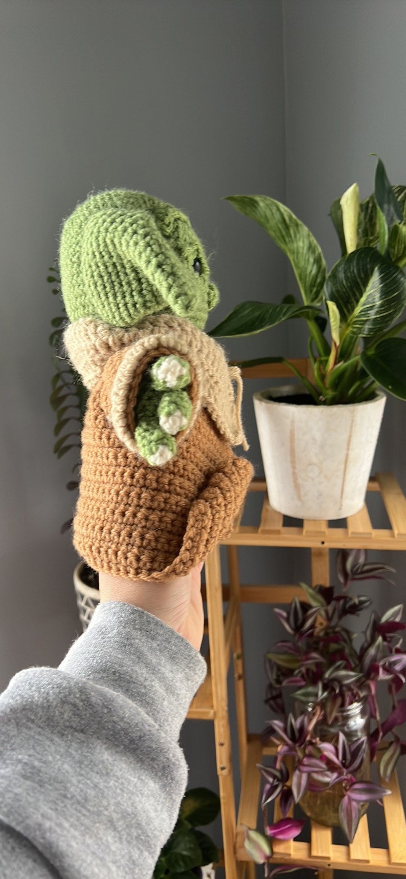 May include: A crocheted green and brown plush toy resembling a character from the Star Wars franchise. The toy is being held by a hand in a grey sweatshirt.