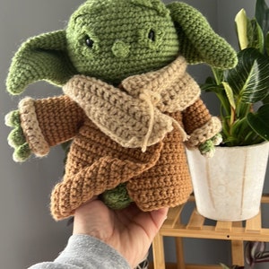 May include: A green crocheted Yoda doll wearing a brown crocheted coat and a beige crocheted scarf. The doll is being held by a hand.