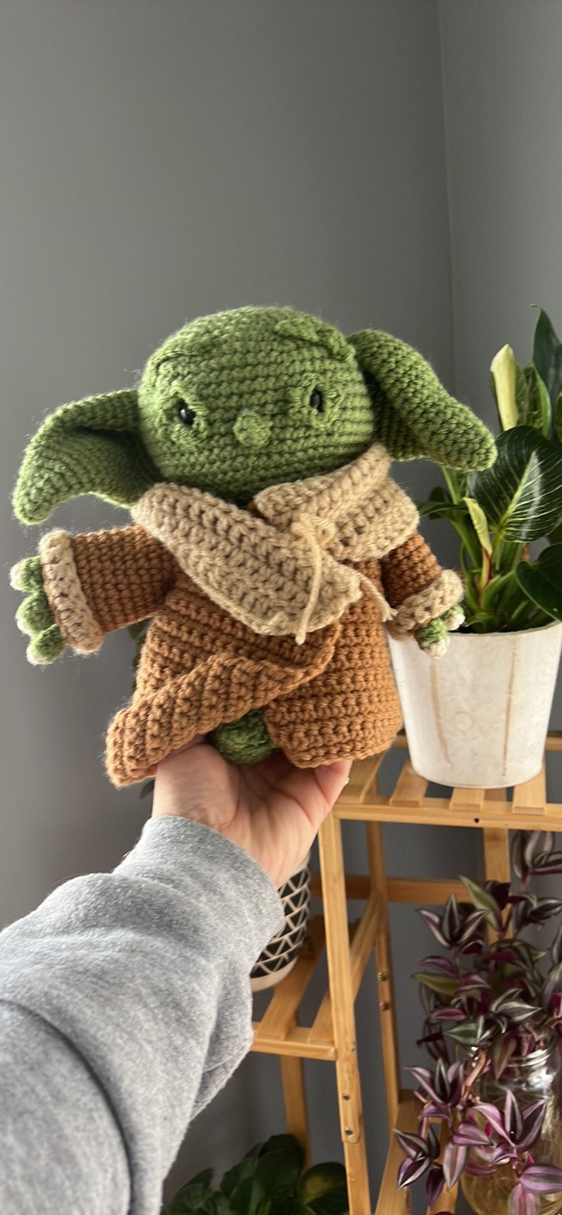 May include: A green crocheted Yoda doll wearing a brown crocheted coat and a beige crocheted scarf. The doll is being held by a hand.