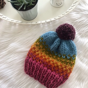 May include: A colourful knitted hat with a pom-pom on top. The hat is striped with pink, orange, yellow, green, and blue yarn.