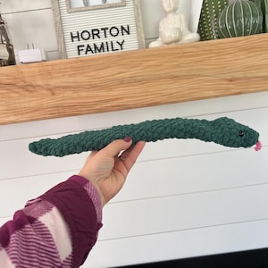 May include: A green crocheted snake toy with a pink nose. The snake is being held by a hand wearing a plaid shirt.
