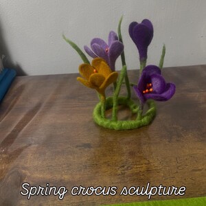 May include: A felt sculpture of a bouquet of purple and yellow crocuses. The flowers are arranged in a circle with green stems and leaves. The sculpture is on a wooden surface with the text "Spring crocus sculpture" below it.