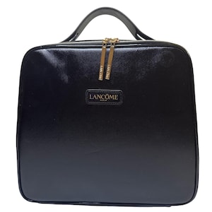 Puede incluir: Un neceser negro y rectangular con asa superior y cremalleras doradas. La funda presenta el logotipo "Lancôme" en letras doradas en la parte delantera. Diseñado para transportar cosméticos y productos de belleza.