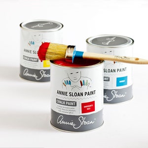 May include: Three cans of Annie Sloan Chalk Paint, one in red, one in blue, and one in white. A paintbrush is resting on top of the red can.