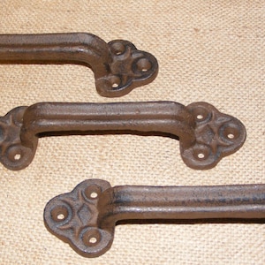 May include: Three rustic, brown cast iron handles. Each handle has a curved grip and decorative ends with multiple holes for mounting. The handles are likely for furniture or cabinetry.
