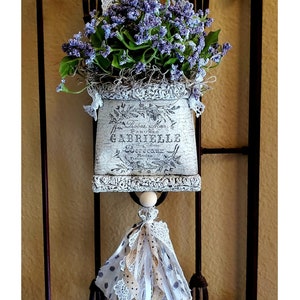 May include: A decorative wall hanging featuring a distressed, off-white planter filled with purple flowers and greenery. The planter has French text and is adorned with lace and a tassel. The piece hangs on a wrought iron gate.