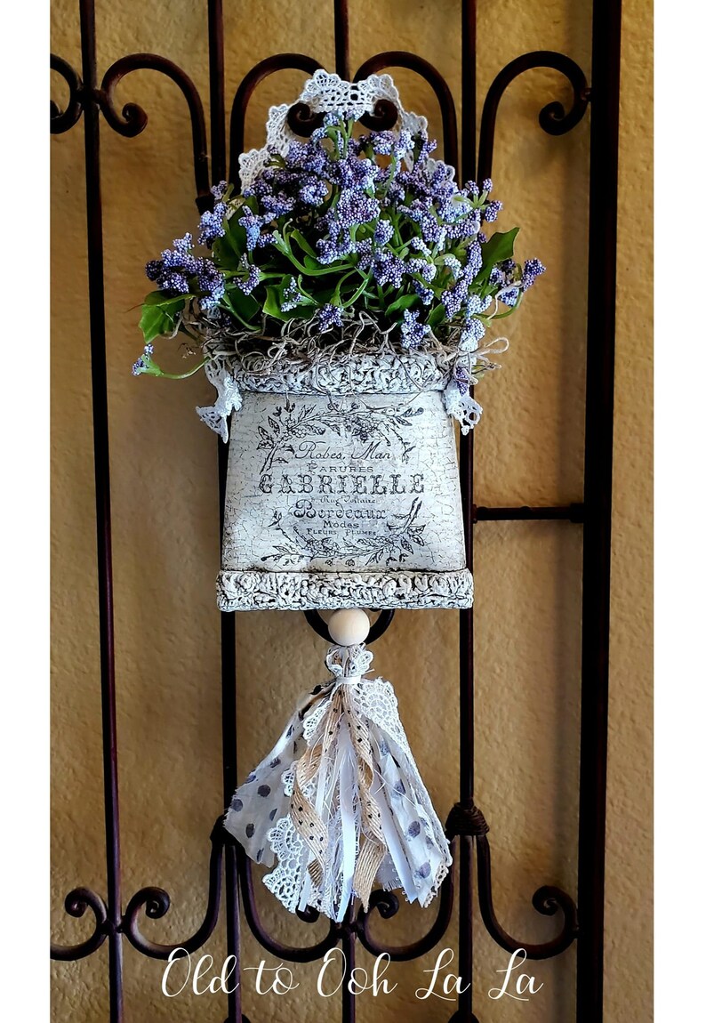 May include: A decorative wall hanging featuring a distressed, off-white planter filled with purple flowers and greenery. The planter has French text and is adorned with lace and a tassel. The piece hangs on a wrought iron gate.
