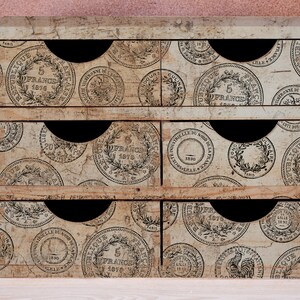 May include: A small, vintage-style wooden storage chest with six drawers. The drawers are decorated with a repeating pattern of antique French coin designs in black and white. The chest has a distressed, aged appearance, with black pull-out handles.