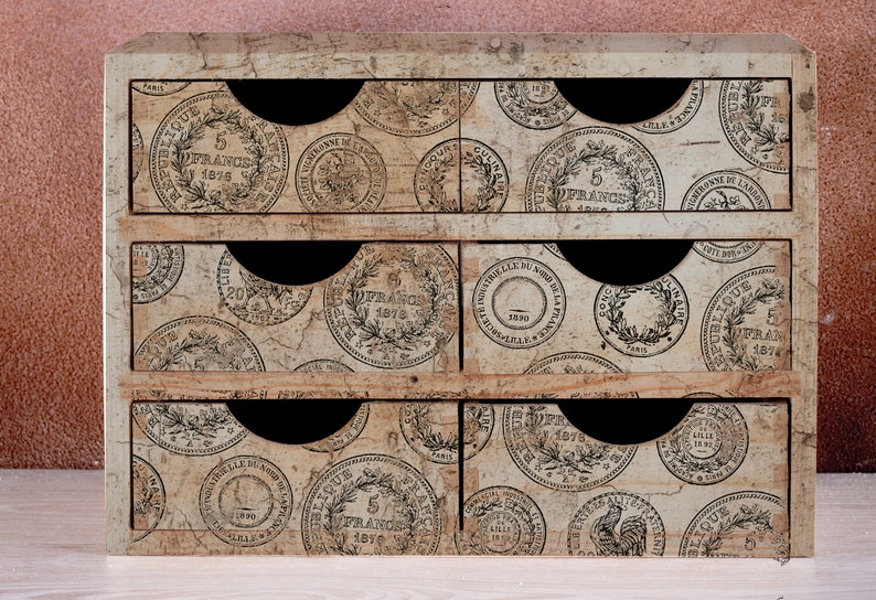 May include: A small, vintage-style wooden storage chest with six drawers. The drawers are decorated with a repeating pattern of antique French coin designs in black and white. The chest has a distressed, aged appearance, with black pull-out handles.