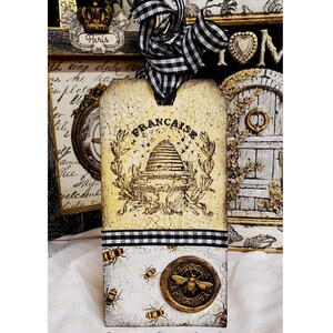 May include: A decorative tag with a French theme, featuring a beehive design and the word "FRANCAISE". The tag is adorned with a black and white checkered ribbon and a gold-colored bee emblem. The bottom section displays bee illustrations. The text "Old to Ooh La La" is at the bottom.