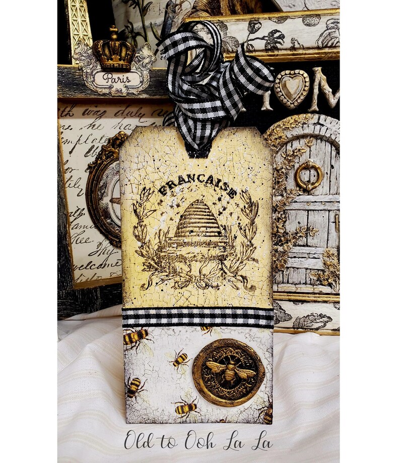 May include: A decorative tag with a French theme, featuring a beehive design and the word "FRANCAISE". The tag is adorned with a black and white checkered ribbon and a gold-colored bee emblem. The bottom section displays bee illustrations. The text "Old to Ooh La La" is at the bottom.