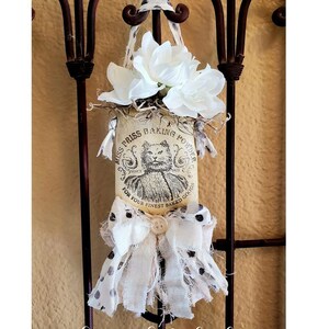 May include: A decorative fabric ornament featuring a vintage-style label with the text "Miss Priss Baking Powder" and a cat illustration. The ornament is adorned with white fabric, a bow, and white flowers. It hangs from a brown metal gate.
