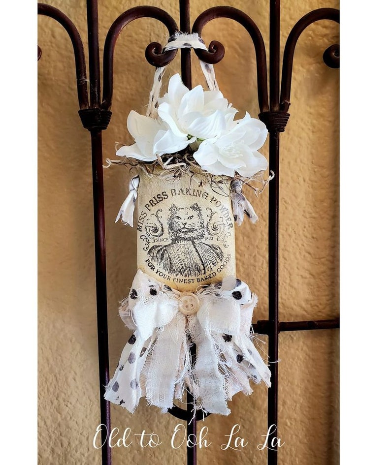 May include: A decorative fabric ornament featuring a vintage-style label with the text "Miss Priss Baking Powder" and a cat illustration. The ornament is adorned with white fabric, a bow, and white flowers. It hangs from a brown metal gate.