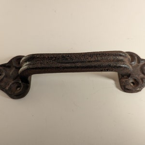 May include: A dark brown cast iron door pull with a decorative design. The pull has a smooth, slightly curved surface and two holes on each side.