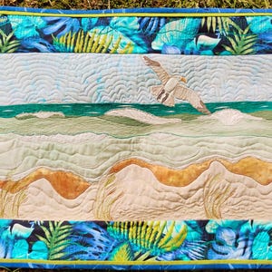 May include: A quilted wall hanging depicting a beach scene with a seagull in flight. The artwork features a sandy beach, ocean waves, and a sky with cloud-like patterns. The border is a tropical leaf pattern in shades of blue and green.