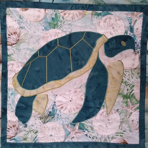May include: A teal blue and beige quilted fabric wall hanging featuring a sea turtle with a detailed shell design. The background is a light blue and white fabric with a pattern of seashells and starfish.