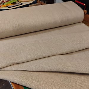 May include: A roll of natural linen fabric, folded in thirds, with a rough, woven texture. The fabric is a light beige color.
