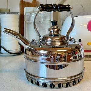 May include: A polished stainless steel stovetop kettle with a cylindrical body, a curved spout, and a black handle. The kettle features a removable lid with a small black knob and a perforated metal base designed for heat distribution on a stove.