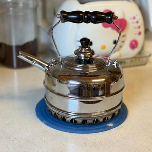 May include: A polished stainless steel stovetop kettle featuring a rounded body, a short spout, and a black wooden handle. The kettle has a removable lid with a small black knob and a circular base with ventilation holes for heat distribution.