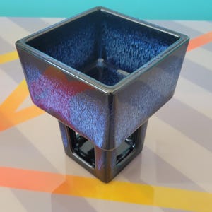 May include: A square ceramic planter with a glossy finish featuring a deep navy blue and black gradient glaze. The upper section is wide and open, while the base narrows into a smaller square pedestal with open cutouts on each of the four sides.