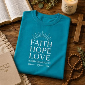 May include: A teal crew neck t-shirt featuring white text that reads FAITH HOPE LOVE 1 CORINTHIANS 13:13, accompanied by a sunburst graphic above the text and an arrow with a small heart in the centre positioned below the scripture reference.