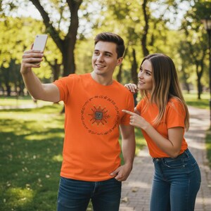 Puede incluir: Dos camisetas naranjas con un diseño gráfico circular. El diseño presenta un cuadrado central con líneas y símbolos radiantes. Las camisetas son usadas por dos personas al aire libre, en un parque.
