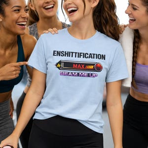 May include: Light blue t-shirt with the text "ENSHITTIFICATION" above a graphic with "MAX" and "BEAM ME UP". The shirt is worn by a person in black leggings, surrounded by smiling people. A water bottle is held in the foreground.